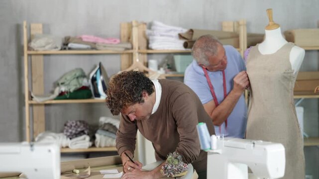 Male clothing designer drawing a sketch of clothes on paper using the client's individual measurements against the background of a tailor in a sewing workshop . High quality 4k footage