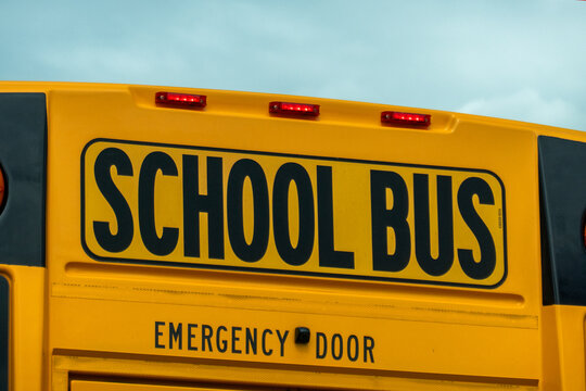 Close up shot of the rear top sign and emergency door warning on a traditional yellow school bus under a cloudy sky.