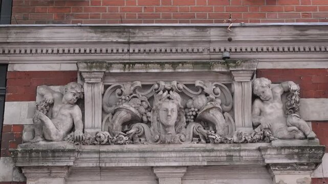 Detailed classical frieze with two reclining figures, central face, and cornucopia above a red brick facade in soft daylight.
