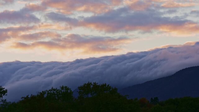 Timelapse of a dramatic sea of clouds flowing over the Jura Mountains above Nyon, Switzerland during sunrise. Low clouds cascade across the ridge beneath colorful morning skies.