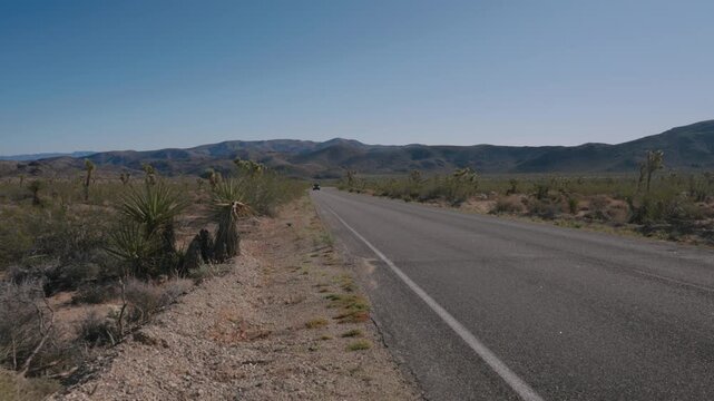 An empty two-lane desert highway stretches toward distant mountains under a clear blue sky.