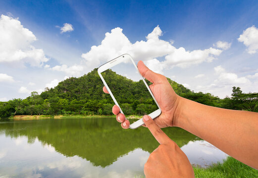 Hand holding smartphone with transparent screen taking photo of beautiful mountain and lake landscape.