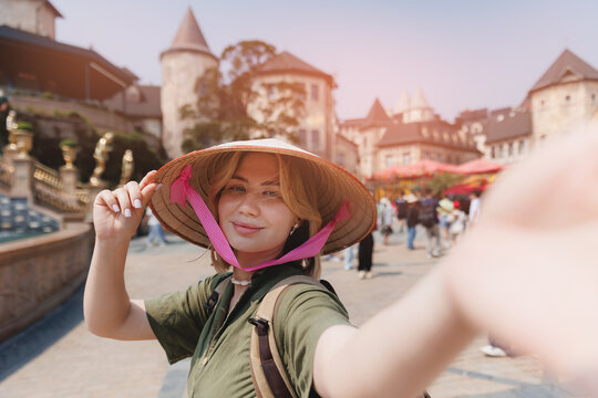 Young caucasian female tourist with backpack making selfie photo at ba na hills, Vietnam wearing traditional conical hat