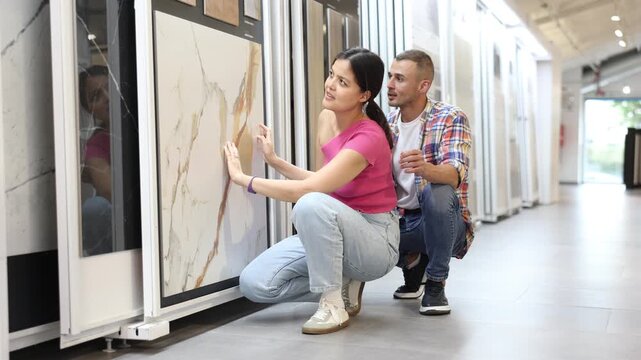 Positive young interracial couple examining technical stone tile samples displayed on sliding exhibit stand in home improvement store, considering colors and textures for renovation project