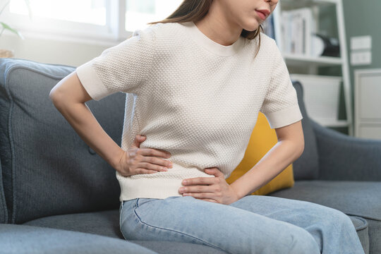 Close-up of a young woman sitting on the sofa and holding her stomach, suffering from stomach ache, abdominal pain or menstrual cramps at home.