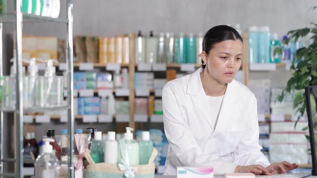 Focused young Asian female pharmacist in white coat using desktop computer to update medicine quantities and manage inventory during routine stock control 