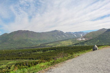 Fototapeta premium Panoramic view of the Chugach Mountains and green valley near Anchorage, Alaska. Summer landscape of Alaskan wilderness with a gravel path, evergreen forest, and mountains with remaining snow patches.