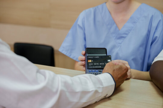 Multiracial patient using credit card to make payment at hospital front desk while caucasian nurse observes during administrative process of medical registration and billing coordination