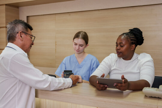 Multiracial female medical staff assisting senior asian male patient at hospital payment counter using credit card reader and digital tablet for healthcare service billing in modern reception area
