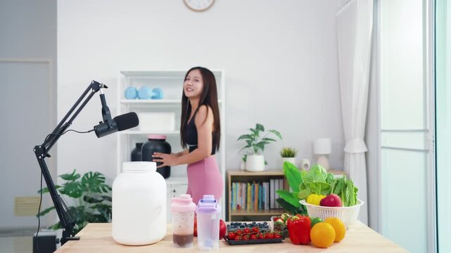 Sporty Asian woman broadcasting vegan lifestyle tips using mobile phone on tripod. Energetic health content creator sharing raw food recipes and fitness motivation online.