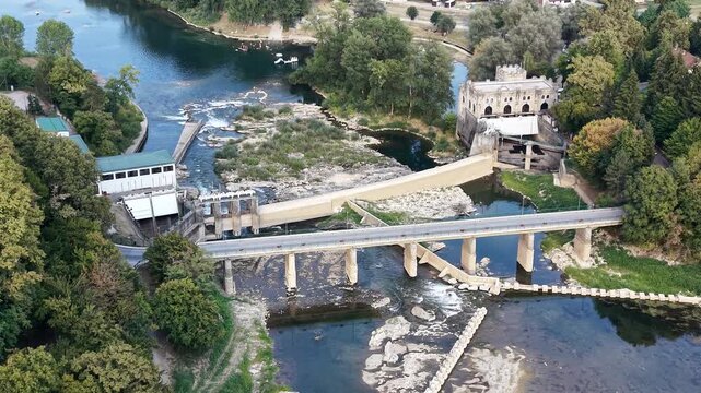 Aerial view of the historic Ozalj Castle and the hydroelectric power plant situated on the Kupa River, surrounded by lush trees and a bridge in Ozalj, Karlovac County, Croatia.