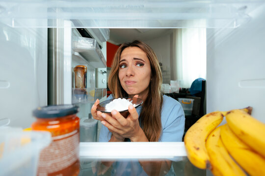 Woman Holding Bowl Of Baking Soda Inside Refrigerator In The Morning. Thoughtful lady holds a small glass bowl filled with baking soda inside an open fridge to remove bad odors