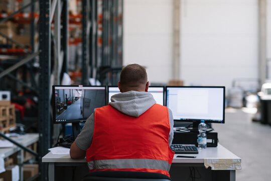 Man in a red reflective vest monitoring stock movements on three computer screens at a warehouse workstation for supply chain and logistics software