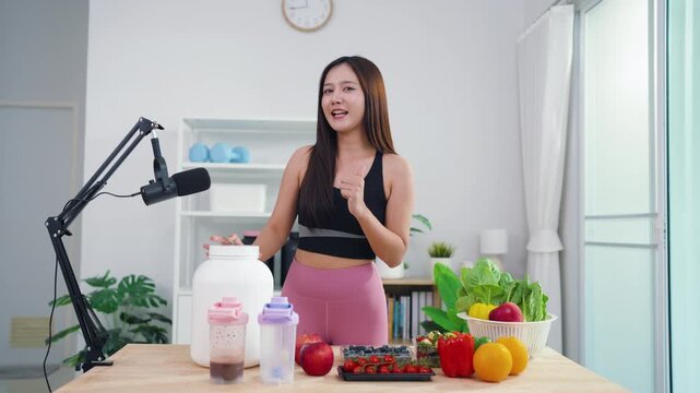 Sporty Asian woman broadcasting vegan lifestyle tips using mobile phone on tripod. Energetic health content creator sharing raw food recipes and fitness motivation online.