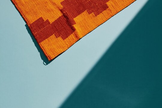 Handwoven kente cloth with bold orange and sienna motifs, featuring a ribbed slubbed texture on a teal surface for cultural heritage and textile design