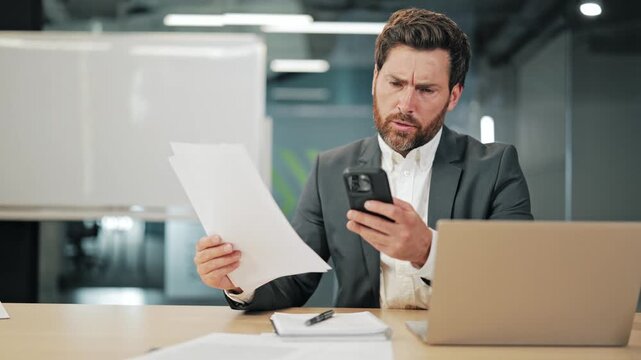 Professional experiencing confusion while comparing documents and smartphone data in modern office environment. Deep in thought over business information, seeking solutions problem solving.