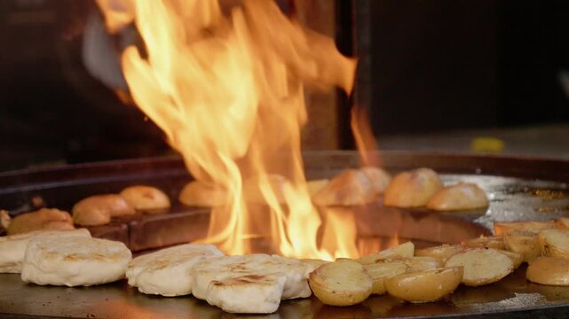 A street food vendor cooks baked potatoes over an open flame at a busy festival. The process shows the sizzling sound and smoky appearance of the food.