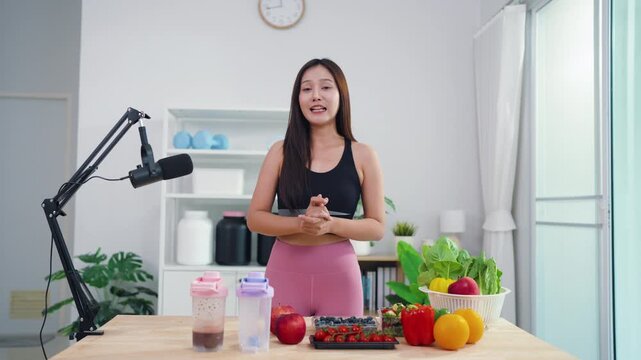 Sporty Asian woman broadcasting vegan lifestyle tips using mobile phone on tripod. Energetic health content creator sharing raw food recipes and fitness motivation online.