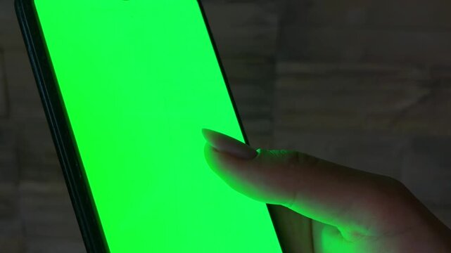 Young woman's hand holding a smartphone with a green screen, her thumb scrolling and tapping the display in a dark room, perfect for compositing custom content on the mobile device