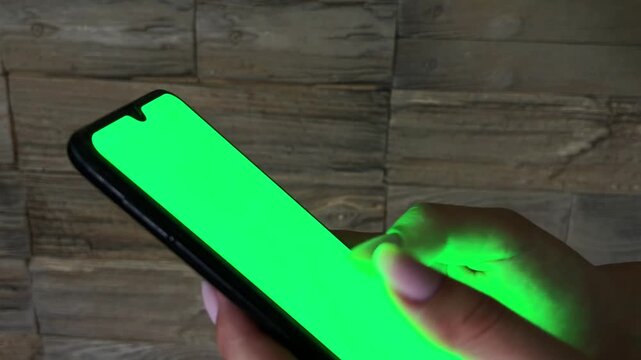 Woman holding a mobile phone with a green screen, scrolling and tapping on the touch display, browsing social media or online content in a dark environment at home