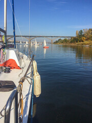 Fototapeta premium Sailing boats on a calm river near an arched bridge, view from the deck of a yacht.