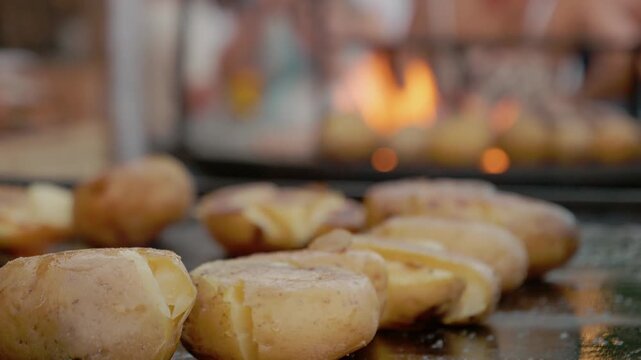 Whole baked potatoes are placed on a grill plate as people gather around at a bustling street food festival. The smell of roasted food fills the air.