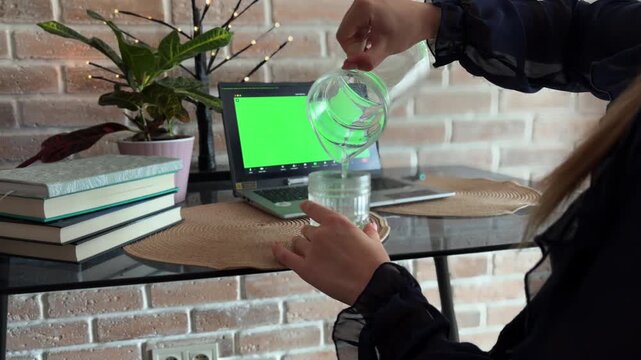 Woman attending online therapy session on a laptop with green screen while pouring water into a glass, expressing her emotions and gesticulating during the meeting