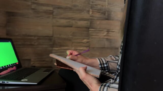 Professional psychotherapist providing online therapy session and taking notes in a notebook while talking with a patient through a laptop with a green screen