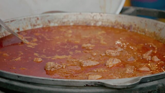 A large cauldron bubbles with meat and tomatoes at a local street food festival. The smell fills the air as people gather around to watch and enjoy the food.