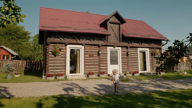 A little boy walks by a traditional house in Bucovina. He stops and looks directly at the camera. The house is made of wood and has a red roof.