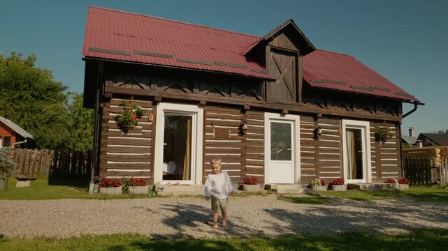 A boy walks barefoot on stones outside a traditional Romanian house in Bucovina. The sunny day highlights the natural beauty around the home and garden.