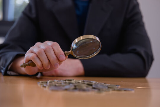 Business professional examining coins with magnifying glass, representing financial analysis, investment research, money management, audit, savings, risk assessment and financial planning.