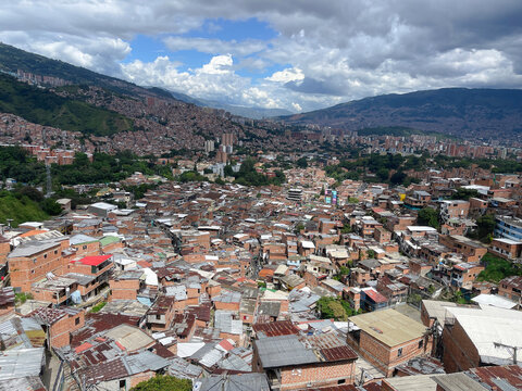 High-angle panoramic view of a densely populated urban hillside favela with brick houses, tin roofs, and distant mountains under clouds.