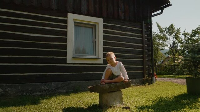 A little boy sits on a stone outside a traditional Romanian house in Bucovina. He looks directly at the camera with a cheerful expression on a sunny day.