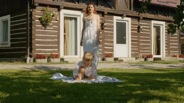 A mother walks towards her child who is sitting on the grass, enjoying some time in front of their traditional house in Bucovina. The house has a wooden structure.