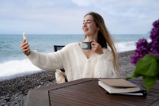 Smiling woman using the front camera of a smartphone to take a selfie by the sea while enjoying a warm drink. Casual travel and digital lifestyle concept