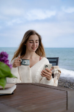Casual portrait of a woman relaxing by the ocean, drinking coffee and using her smartphone. Modern seaside lifestyle and leisure concept.