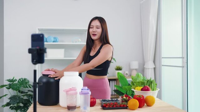 Female fitness coach filming post-workout nutrition guide with fresh green veggies. Smiling Asian girl recording healthy smoothie preparation for wellness social media channel.
