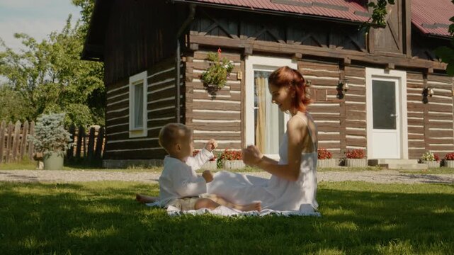 A mother enjoys time with her child while sitting on grass outside a traditional house in Bucovina. They share laughs and play with joy under the sun.