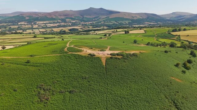 Drone flying low above lush green bracken and agricultural fields in summer weather, distant ridges of Pen y Fan rising under clear skies in Bannau Brycheiniog