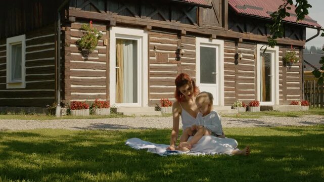 A mother sits on the grass with her child and reads a book. They are in front of a traditional house in Bucovina. It is a sunny day with a clear sky.