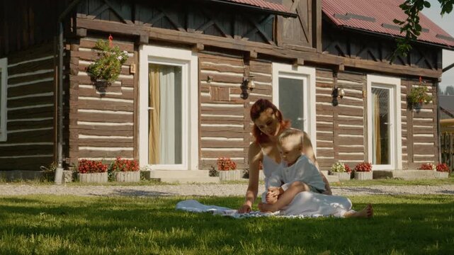 A mother sits on the grass with her child in Bucovina. They enjoy a story together outside a traditional house, surrounded by nature and the warmth of the sun.