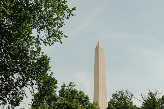 Washington Monument rises above green trees beneath a hazy summer sky.