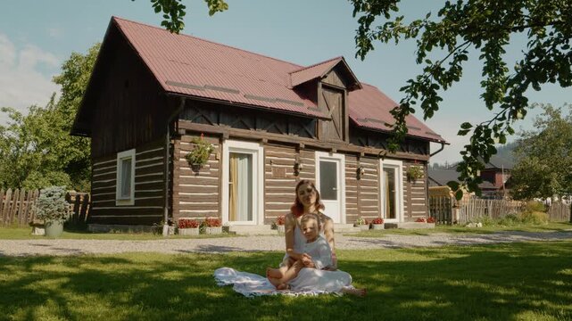 A woman and her son sit together on the grass, looking at the camera. Behind them is a traditional house in Bucovina. The day is sunny and bright.