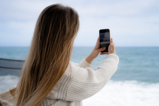 Rear view of a woman using a smartphone to photograph the sea during a cloudy day. Travel photography, social media, and the modern seaside lifestyle.