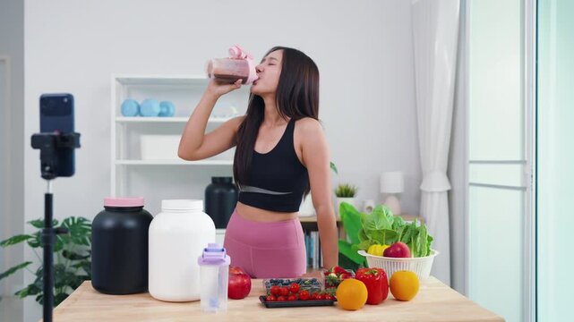 Female fitness coach filming post-workout nutrition guide with fresh green veggies. Smiling Asian girl recording healthy smoothie preparation for wellness social media channel.