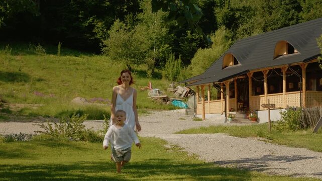 A woman and her boy run together in the grass near a traditional house in Bucovina. The sun shines and the greenery surrounds them as they enjoy their time outside.