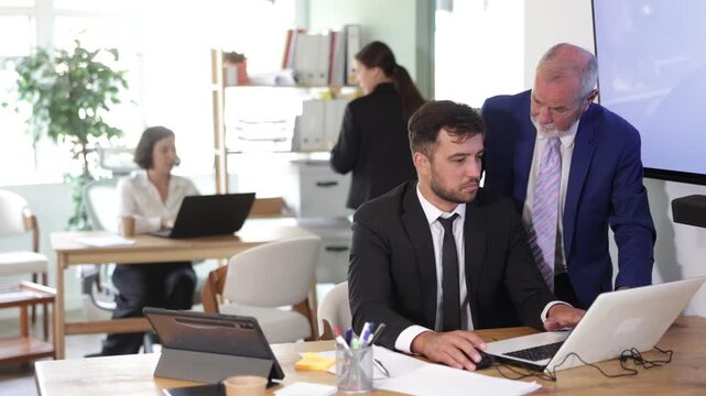 Senior employee of the office bank stands next to the male employee and helps him deal with electronic invoices on his laptop. Experienced office employee explains work tasks. High quality 4k footage