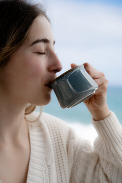 Close-up of a woman savoring a warm drink outdoors in nature beside the sea. Mindful living, relaxation and simple pleasures in a coastal setting.