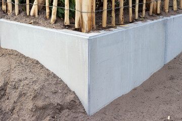 Concrete retaining wall corner with sandy soil and wooden fence posts supporting vegetation in a landscaped garden setting © Yahor